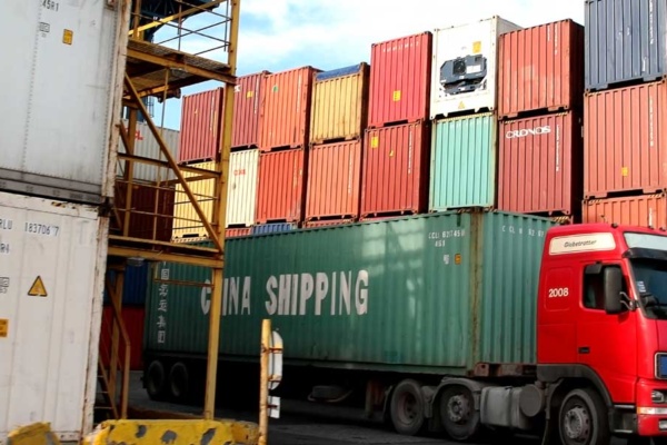 A truck carrying a China Shipping container in front of a stack of shipping containers