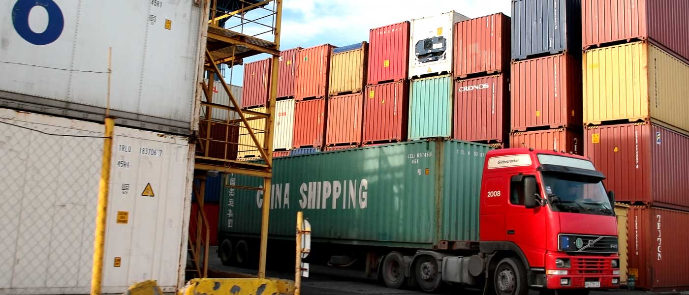 A truck carrying a China Shipping container in front of a stack of shipping containers